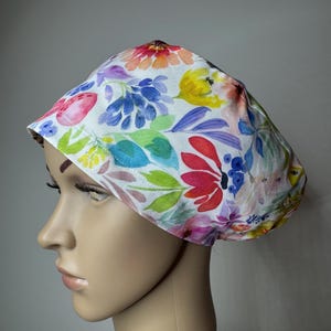 May include: A scrub cap featuring a white background with a vibrant watercolour floral print in shades of red, yellow, blue, pink, and green. The fabric head covering has a rounded crown and a wide, stitched band that fits snugly around the head.