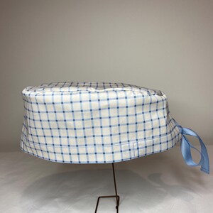 Può includere: Un cappello da chirurgo a quadri bianchi e blu con un nastro azzurro chiaro. Il cappello è in tessuto ed è esposto su un supporto metallico. Il motivo a quadri è una griglia di linee azzurre chiare su sfondo bianco.