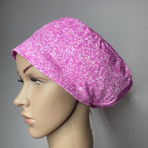 May include: A scrub cap featuring a vibrant pink and white speckled pattern. The fabric has a dense, multi-tonal design with small white and light pink dots scattered across a bright magenta background, creating a textured visual effect.