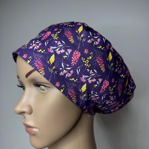 May include: A scrub cap featuring a deep purple background with a repeating pattern of small pink, yellow, and white wildflowers, thin green stems, and tiny bees. The cap has a rounded, gathered crown and a wide, fitted band around the forehead.