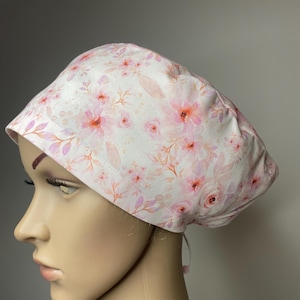 May include: A scrub cap featuring a white background with a soft watercolour floral pattern in shades of pink, peach, and light purple. The fabric head covering has a rounded crown and a wide, stitched band around the base with a tie closure.