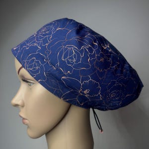 May include: A navy blue scrub cap featuring a repeating pattern of rose and daffodil outlines in a metallic copper colour. The cap has a rounded crown and a black drawstring cord with a small bead at the back for an adjustable fit.