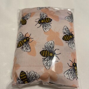 May include: A rectangular fabric pouch encased in clear plastic packaging. The fabric features a repeating pattern of detailed honeybees with yellow and black striped bodies and white wings, set against a light pink background with faint peach-colored butterfly silhouettes.