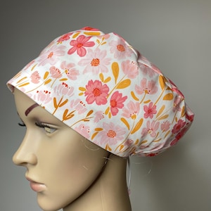Può includere: Un cappuccio chirurgico bianco con un motivo floreale rosa. Il cappuccio presenta un motivo ripetuto di fiori rosa e corallo con foglie giallo-arancio. Il cappuccio è legato sul retro con cordini bianchi.