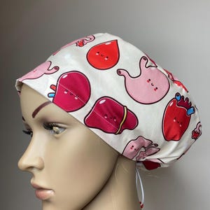 以下が含まれることがあります： ピンクと赤の漫画の臓器がプリントされた白いスクラブハット。臓器は笑顔で、心臓、胃、肝臓、血のしずくが含まれています。