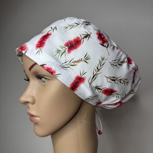 May include: A white scrub cap featuring a repeating botanical print of red bottlebrush flowers and green foliage. The cap has a rounded crown, a wide front band, and a tie-back closure with small red beads at the ends of the white fabric ties.