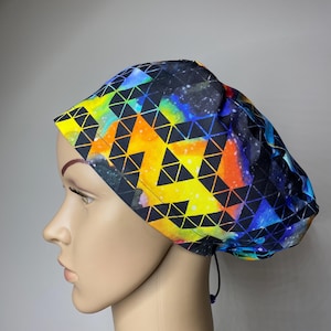 Può includere: Una cuffia da sala operatoria con una vivace stampa ispirata alle galassie e una sovrapposizione geometrica di triangoli neri. Il tessuto mostra uno spettro di tonalità brillanti giallo, arancione, blu e viola su sfondo scuro, con cordino regolabile.