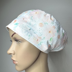 May include: A scrub cap featuring a white background with a soft watercolour floral pattern in shades of pale pink, peach, and light green. The fabric hat has a rounded crown and a wide, structured band that fits snugly around the head.