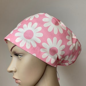 May include: Pink surgical scrub hat with a white daisy print. The hat has a drawstring tie.