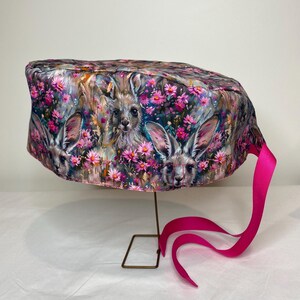 May include: A scrub cap featuring a colorful print of kangaroos amidst pink flowers. The cap has a pink tie and is displayed on a gold-colored wire stand. The fabric has a watercolor effect.