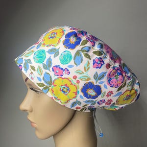 May include: A scrub cap featuring a light pink background with a vibrant floral pattern in shades of yellow, blue, pink, and green. The cap has a rounded crown and includes a white adjustable drawstring cord with a small blue plastic toggle.