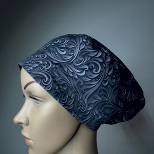 May include: A dark charcoal grey scrub cap featuring a tonal, embossed-style swirling paisley pattern. The head covering has a fitted band across the forehead and a rounded, gathered back section that provides a secure and comfortable fit.