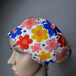 May include: A scrub cap featuring a cream-coloured background with a repeating pattern of large, stylised flowers in shades of pink, yellow, red, and blue. The design includes small white dots on the flower centres and blue paint-like splatters.