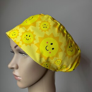 May include: A yellow scrub cap featuring a repeating pattern of bright yellow suns with smiling faces. Each sun has black eyes, a curved black mouth, and small pink cheeks. The fabric is gathered at the back with a stitched hem along the front edge.