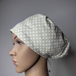 May include: A scrub cap featuring a light beige fabric base with a repeating pattern of small white polka dots. The cap has a rounded, gathered design with a wide front band and a thin adjustable tie closure visible at the back of the head.