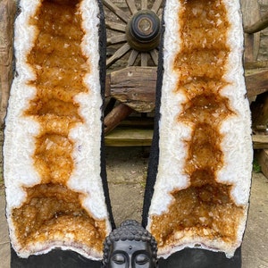 Op de afbeelding: Twee grote, natuurlijke geodekristallen met een witte buitenkant en een goudbruine binnenkant. De kristallen staan ​​rechtop op een stenen oppervlak. Een zwart Boeddhabeeld staat voor de kristallen.