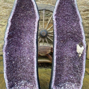 Op de afbeelding: Twee grote amethistgeoden, elk met een diep paarse binnenkant en een zwarte buitenkant. De geoden staan ​​rechtop op een stenen oppervlak.