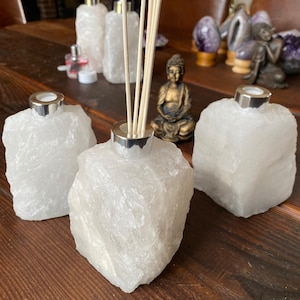 Può includere: Tre bottiglie diffusori in cristallo bianco con tappi argentati e bastoncini di legno. Le bottiglie sono su una superficie di legno con una piccola statua dorata di Buddha sullo sfondo.