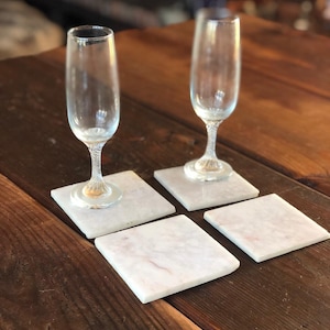 Op de afbeelding: Vier witte marmeren onderzetters met een roze aderingspatroon, twee champagnefluiten staan op de onderzetters op een houten tafel.