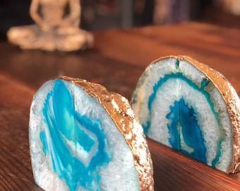 Puede incluir: Dos sujetalibros hechos de ágata con ribete dorado. La ágata es blanca con remolinos azules y verdes. Los sujetalibros están sobre una superficie de madera.