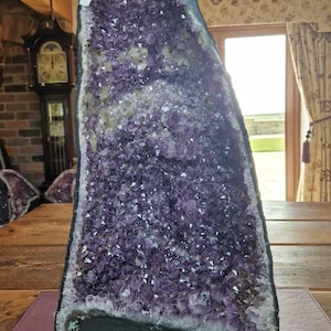 Op de afbeelding: Een grote amethistgeode met een paarse en witte kristallenformatie. De geode staat op een houten tafel met een paarse mat.