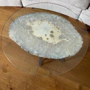 May include: A round glass coffee table with a large, natural agate slice as the tabletop. The agate is white with brown and gray markings. The table has a black metal base.