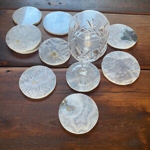 Op de afbeelding: Een set van negen witte agaat-onderzetters met verschillende patronen en een helder glazen wijnglas op een houten oppervlak.
