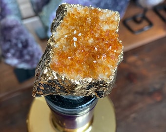 Puede incluir: Un tapón de botella chapado en oro con un gran cristal de citrino naranja en la parte superior. El cristal es rugoso y tiene una forma natural y orgánica.