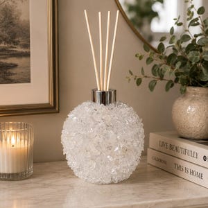 Könnte beinhalten: Ein kugelförmiger Glas-Raumduftdiffusor, der mit klaren, strukturierten, kristallartigen Splittern bedeckt ist. Der Diffusor hat einen silberfarbenen Metallkragen an der Oberseite und vier helle Holzstäbchen, die aus der Öffnung ragen.