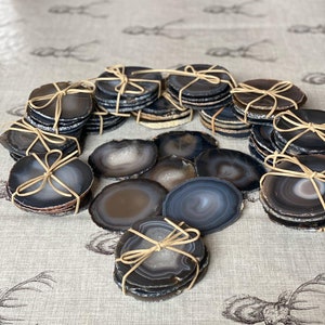 May include: A set of agate coasters, each with a unique pattern of black, grey, and white. The coasters are tied together with twine and are arranged in a circle on a light brown surface.