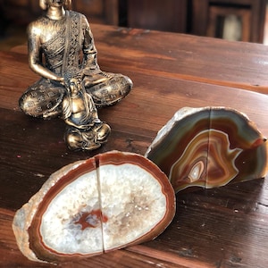 May include: Two agate bookends with a natural crystal pattern. The bookends are in shades of brown, white, and orange. A gold-coloured statue of a meditating Buddha sits on a wooden surface.
