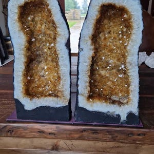 Op de afbeelding: Twee grote, natuurlijke geode-kristalformaties met een zwarte basis. De kristallen zijn lichtgeel van kleur en zijn gerangschikt in een cluster. De geode is open en de kristallen zijn zichtbaar van binnen. De geode staat op een houten oppervlak.