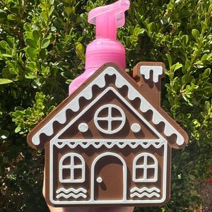 May include: A brown and white gingerbread house-shaped soap dispenser with a pink pump. The house has a door, windows, and a chimney. The dispenser is held in front of a green leafy background.