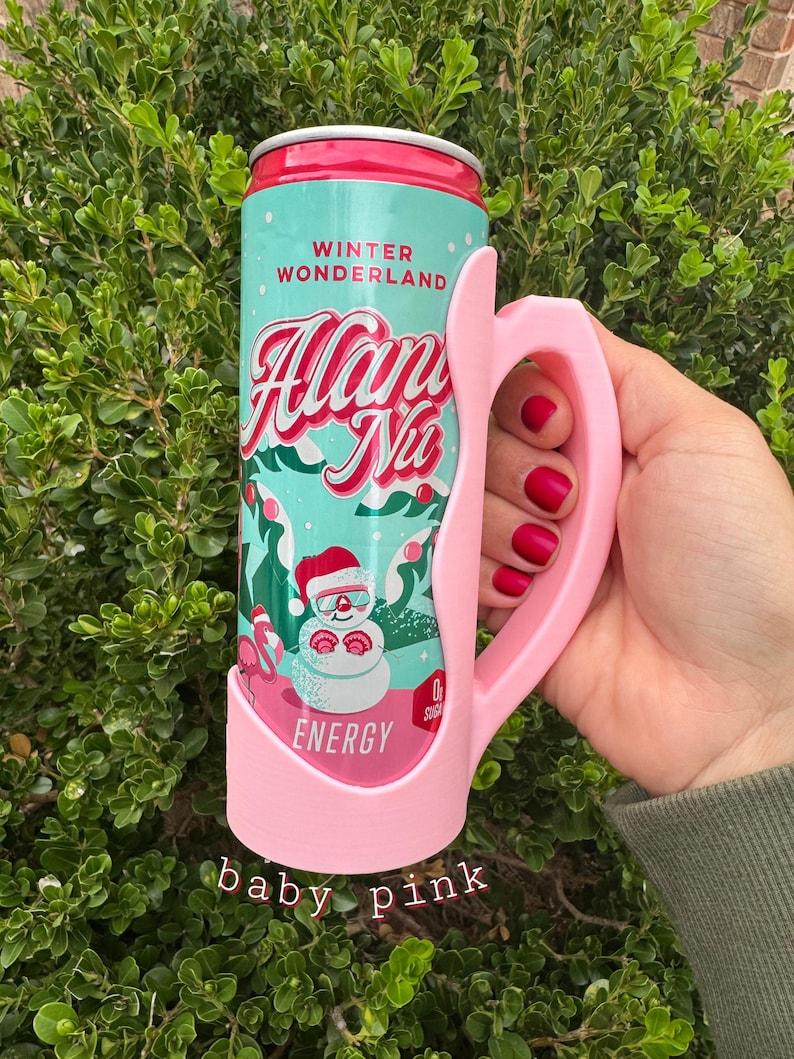 K&ouml;nnte beinhalten: Ein babyrosa Dosenhalter mit Griff, der eine Dose Energy-Drink "Winter Wonderland" h&auml;lt. Die Dose zeigt eine Schneemann-Grafik und die Worte "Alan Nu". Der Halter wird von einer Hand mit rotem Nagellack gehalten.