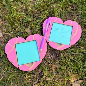 May include: Two heart-shaped memo holders with a light pink and yellow design, each holding a square, light blue sticky note. One note has the text "Snowy made it!" and a heart symbol. A purple pen rests between the holders.