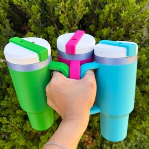May include: Three colorful travel mugs in green, pink, and blue are held up against a green leafy background. Each mug has a white lid with a colored slider and a matching handle. The mugs are cylindrical with a tapered base.