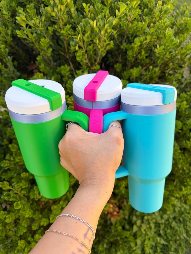 May include: Three colorful travel mugs in green, pink, and blue are held up against a green leafy background. Each mug has a white lid with a colored slider and a matching handle. The mugs are cylindrical with a tapered base.