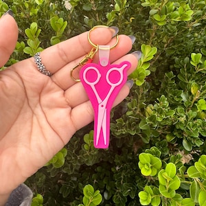 May include: A pink scissor-shaped keychain with a gold-colored keyring and clasp. The scissors are a lighter pink color and are set against a bright pink background. The keychain is held in front of a green leafy background.