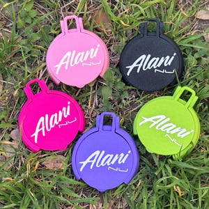May include: Five colorful, round accessories with a handle, resting on grass. Each accessory features the word "Alani" in white script. Colors include pink, black, hot pink, purple, and lime green.