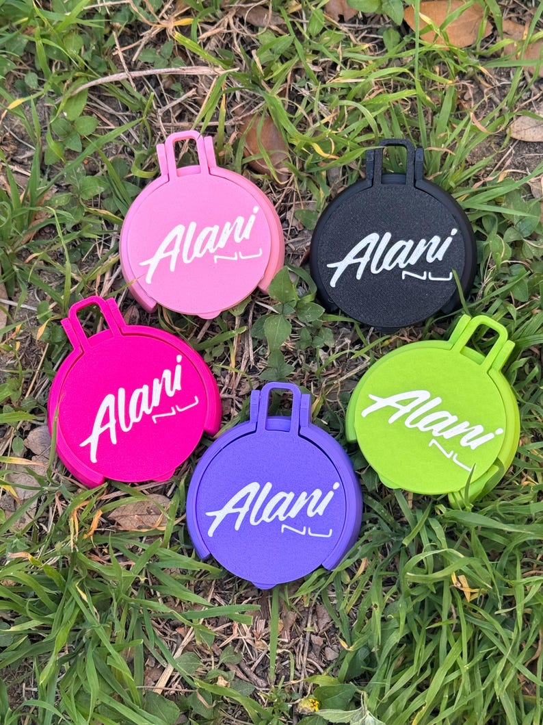 May include: Five colorful, round accessories with a handle, resting on grass. Each accessory features the word "Alani" in white script. Colors include pink, black, hot pink, purple, and lime green.