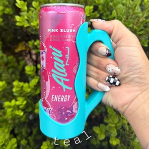 K&ouml;nnte beinhalten: Eine rosa Alani Nu Energy-Drink-Dose, die in einem t&uuml;rkisfarbenen Griff gehalten wird. Die Dose ist mit "Pink Slush" beschriftet und hat ein Erdbeer-Design. Der t&uuml;rkisfarbene Griff ist so konzipiert, dass er eng an die Dose passt.