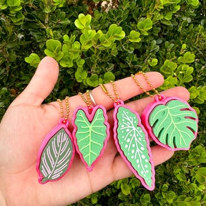 May include: A collection of five leaf-shaped keychains. Each keychain features a different green leaf design with pink borders and gold chain attachments. The leaves vary in shape and detail, showcasing a variety of plant species.