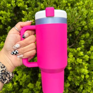 May include: A vibrant pink tumbler with a handle is held against a backdrop of green foliage. The tumbler features a white and silver lid with a pink closure. The handle matches the tumbler's bright pink color. This insulated drinkware is designed for cold beverages.
