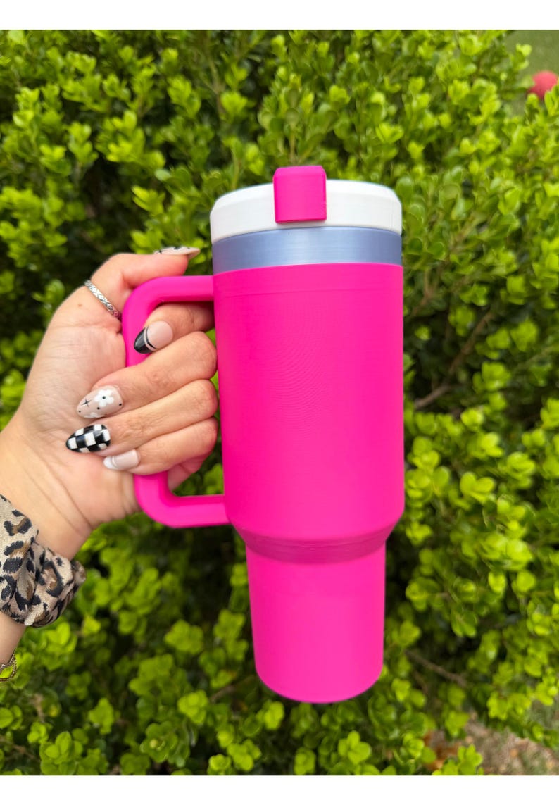 May include: A vibrant pink tumbler with a handle is held against a backdrop of green foliage. The tumbler features a white and silver lid with a pink closure. The handle matches the tumbler's bright pink color. This insulated drinkware is designed for cold beverages.