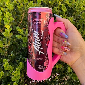 K&ouml;nnte beinhalten: Eine rosa Dose Alani Nu Cherry Bomb Energy-Drink in einem passenden rosa Dosenhalter. Die Dose hat einen dunkelroten Hintergrund mit wei&szlig;em Text und Grafiken. Der Dosenhalter ist mit "peony pink" beschriftet.