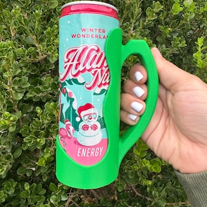 K&ouml;nnte beinhalten: Ein gr&uuml;ner 3D-gedruckter Dosenhalter mit Griff, der eine Dose Energy-Drink "Allan Na" h&auml;lt. Die Dose hat ein Winterwunderland-Design mit einem Schneemann und einem Flamingo. Die Dose hat einen roten Deckel und einen t&uuml;rkisfarbenen K&ouml;rper.