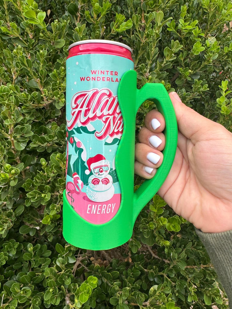 K&ouml;nnte beinhalten: Ein gr&uuml;ner 3D-gedruckter Dosenhalter mit Griff, der eine Dose Energy-Drink "Allan Na" h&auml;lt. Die Dose hat ein Winterwunderland-Design mit einem Schneemann und einem Flamingo. Die Dose hat einen roten Deckel und einen t&uuml;rkisfarbenen K&ouml;rper.
