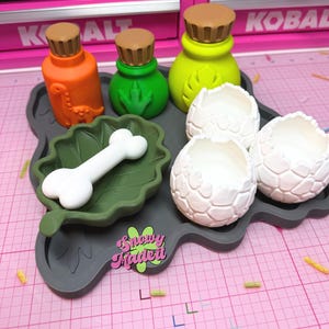 May include: A set of three colorful bottles with brown caps, a white bone on a dark green leaf-shaped tray, and three white textured egg-shaped containers. The tray features a pink and green logo with the text Snowy Madeit.