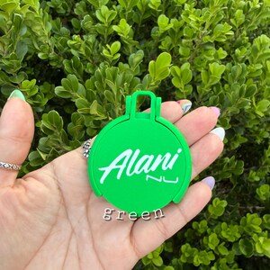May include: A bright green, circular item with the name "Alani" in white script. The word "green" is printed below the name. The item has a handle-like design at the top and is held in a hand against a backdrop of green foliage.