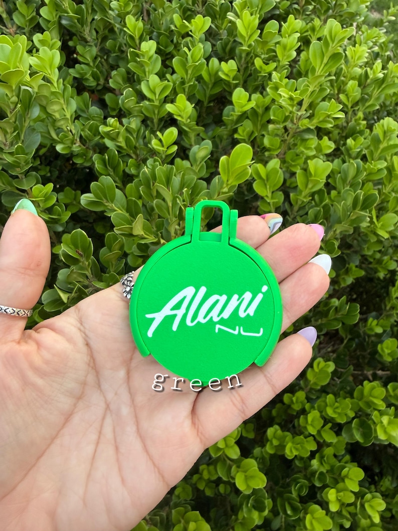 May include: A bright green, circular item with the name "Alani" in white script. The word "green" is printed below the name. The item has a handle-like design at the top and is held in a hand against a backdrop of green foliage.